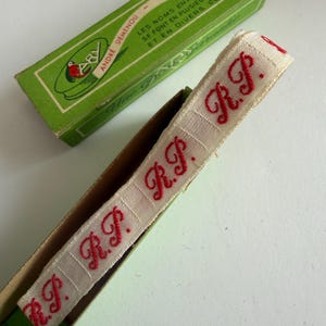 May include: A beige woven fabric ribbon featuring the repeating red embroidered letters R.P. in a cursive script. The ribbon is partially contained within a rectangular green cardboard box labeled Andr&eacute; Demenou with French text.