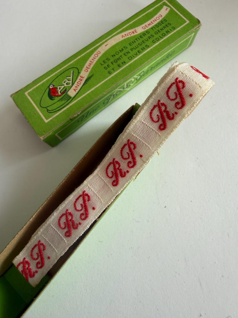 May include: A beige woven fabric ribbon featuring the repeating red embroidered letters R.P. in a cursive script. The ribbon is partially contained within a rectangular green cardboard box labeled Andr&eacute; Demenou with French text.