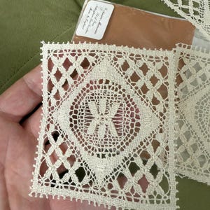 May include: A delicate white lace doily with a square shape and a central floral design. The doily is handmade and has a fine, intricate pattern.