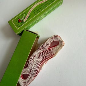 May include: A roll of white fabric tape with red lettering inside a green cardboard box. The box features the text ANDRE DEMENOU, LES NOMS ENTIERS TISSES SE FONT EN PLUSIEURS GENRES ET EN DIVERS COLORIS, and the script text Une Qualit&eacute; Garantie.
