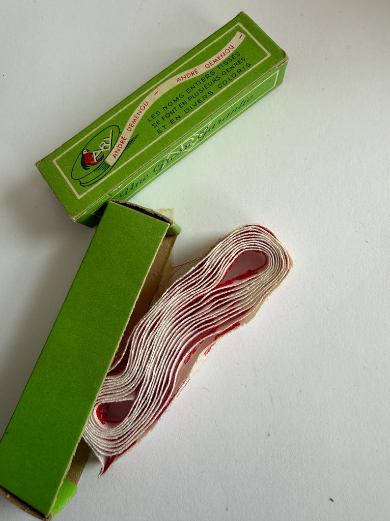 May include: A roll of white fabric tape with red lettering inside a green cardboard box. The box features the text ANDRE DEMENOU, LES NOMS ENTIERS TISSES SE FONT EN PLUSIEURS GENRES ET EN DIVERS COLORIS, and the script text Une Qualit&eacute; Garantie.