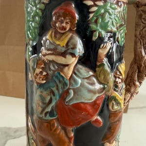 May include: A ceramic beer stein featuring a high-relief, multi-coloured scene of figures in a forest setting. The design includes a person wearing a red hat and a blue top, with green foliage and brown tree-like textures on the handle and rim.