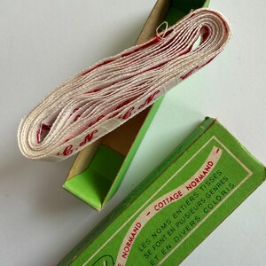 May include: A roll of white woven fabric tape with red cursive lettering reading C.N. resting inside a green cardboard box. The box lid features the text COTTAGE NORMAND and LES NOMS ENTIERS TISS&Eacute;S SE FONT EN PLUSIEURS GENRES ET EN DIVERS COLORIS.