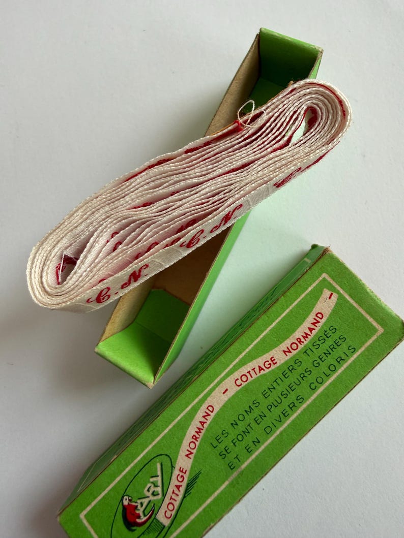 May include: A roll of white woven fabric tape with red cursive lettering reading C.N. resting inside a green cardboard box. The box lid features the text COTTAGE NORMAND and LES NOMS ENTIERS TISS&Eacute;S SE FONT EN PLUSIEURS GENRES ET EN DIVERS COLORIS.