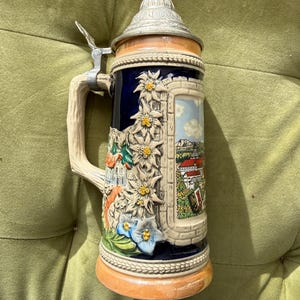 Peut inclure: Une chope à bière en céramique avec un couvercle métallique muni d'un levier. Le corps est bleu foncé avec un relief de fleurs d'edelweiss blanches, de feuillage vert et une scène paysagère peinte montrant des bâtiments et des montagnes.