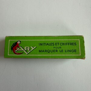 May include: A rectangular green box featuring a parrot illustration and the text ARY INITIALES ET CHIFFRES POUR MARQUER LE LINGE. The packaging has a white border and black lettering, presenting a vintage design for marking fabric items.
