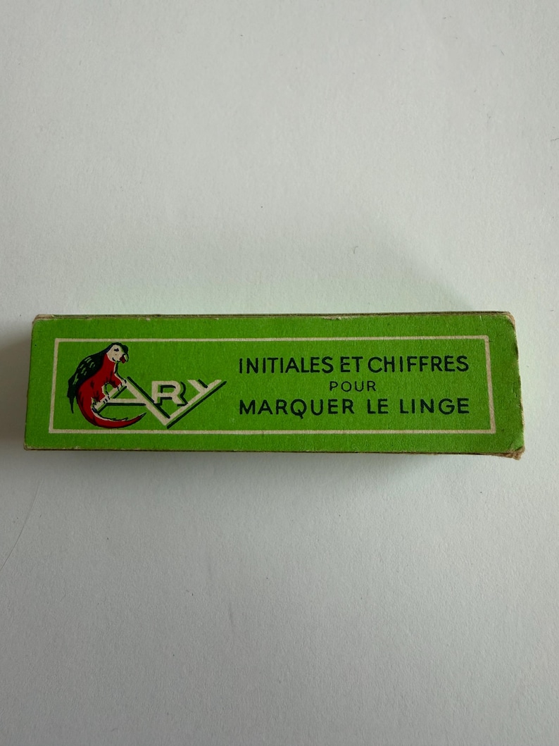 May include: A rectangular green box featuring a parrot illustration and the text ARY INITIALES ET CHIFFRES POUR MARQUER LE LINGE. The packaging has a white border and black lettering, presenting a vintage design for marking fabric items.