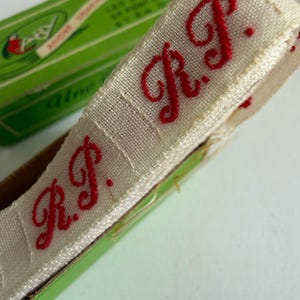 May include: A roll of off-white woven fabric tape featuring the embroidered red initials R.P. in a cursive script. The tape is partially contained within a green cardboard box with visible text, showcasing a repeating pattern of the red monogram.