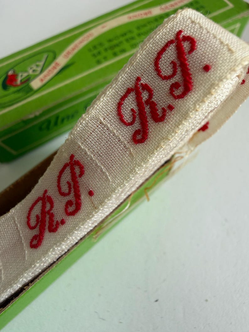 May include: A roll of off-white woven fabric tape featuring the embroidered red initials R.P. in a cursive script. The tape is partially contained within a green cardboard box with visible text, showcasing a repeating pattern of the red monogram.