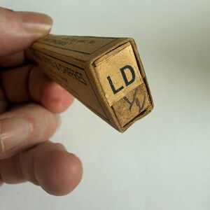 May include: A rectangular tan cardboard box with black text on the side and the letters LD printed in black on the end flap. Below the letters LD, a handwritten symbol resembling a fraction or a mark is visible on a small piece of tape.