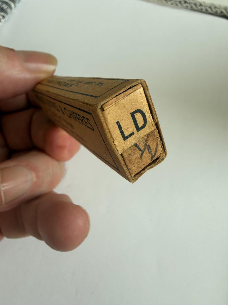 May include: A rectangular tan cardboard box with black text on the side and the letters LD printed in black on the end flap. Below the letters LD, a handwritten symbol resembling a fraction or a mark is visible on a small piece of tape.