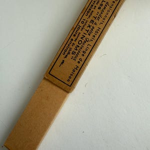 May include: A rectangular tan cardboard box with a sliding sleeve. The sleeve features black printed French text that reads: Pour Pensionnats, H&ocirc;tels, Linge de Maison, demandez &agrave; votre fournisseur Les TEXTINOMS, Noms tiss&eacute;s en entier, initiales suivies de chiffres, etc., Livraison 10 jours environ.
