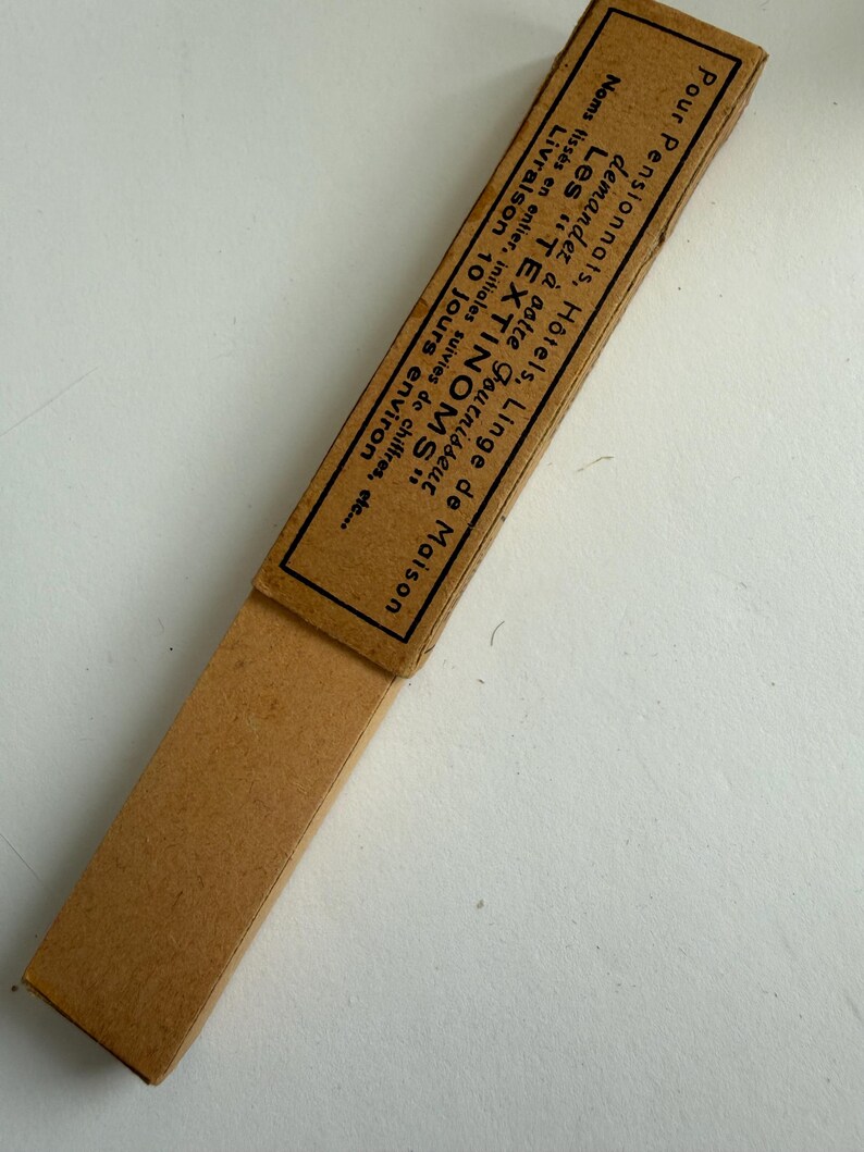 May include: A rectangular tan cardboard box with a sliding sleeve. The sleeve features black printed French text that reads: Pour Pensionnats, H&ocirc;tels, Linge de Maison, demandez &agrave; votre fournisseur Les TEXTINOMS, Noms tiss&eacute;s en entier, initiales suivies de chiffres, etc., Livraison 10 jours environ.