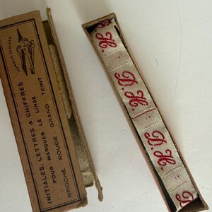 May include: A beige fabric ribbon with red embroidered initials H.G. inside a rectangular cardboard box. The box lid features black text reading INITIALES, LETTRES & CHIFFRES POUR MARQUER LE LINGE BROCH&Eacute; ROUGE GRAND TEINT and a small graphic.