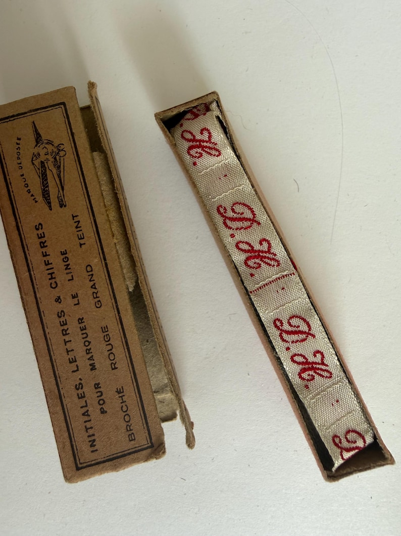 May include: A beige fabric ribbon with red embroidered initials H.G. inside a rectangular cardboard box. The box lid features black text reading INITIALES, LETTRES & CHIFFRES POUR MARQUER LE LINGE BROCH&Eacute; ROUGE GRAND TEINT and a small graphic.