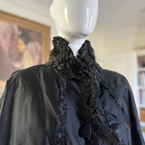 May include: A black cape with a high, ruffled collar and decorative trim. The cape features intricate lace and ribbon details, creating a vintage aesthetic. The garment is displayed on a mannequin.