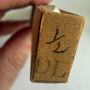May include: A rectangular brown cardboard box with a textured surface. The end panel features the handwritten fraction 1/2 in black ink above the printed letters DL. A small portion of white fabric is visible peeking out from the top edge.