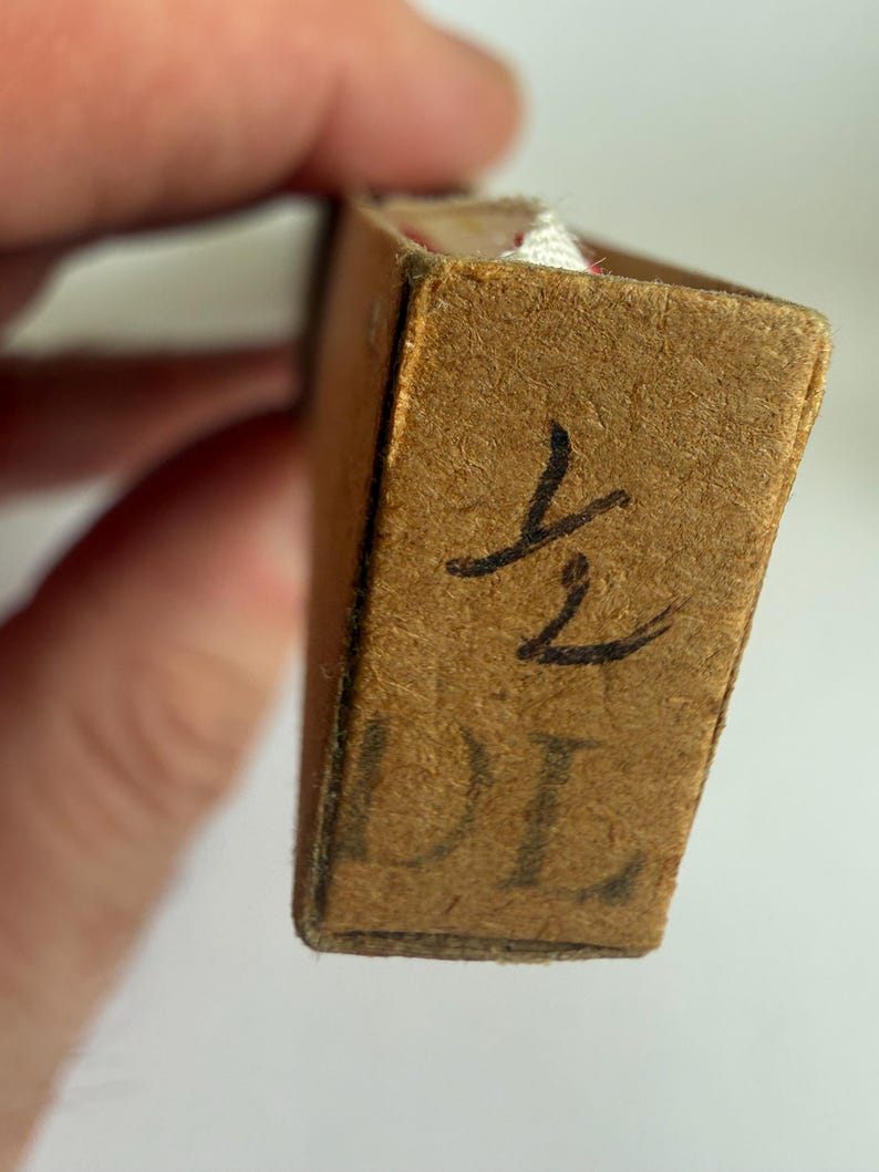 May include: A rectangular brown cardboard box with a textured surface. The end panel features the handwritten fraction 1/2 in black ink above the printed letters DL. A small portion of white fabric is visible peeking out from the top edge.