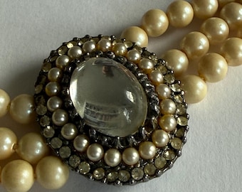 May include: A multi-strand necklace featuring cream-colored round beads and a central oval brooch. The brooch contains a large, clear, smooth cabochon stone surrounded by a border of small pearls and a outer ring of tiny, clear, faceted crystals.