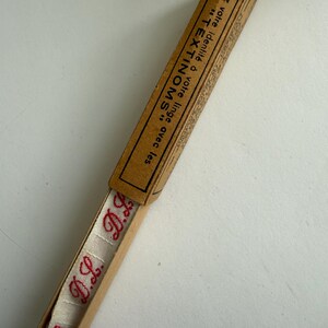 May include: A long, narrow, light brown cardboard box containing a strip of off-white fabric tape with the red embroidered initials D.L. The box lid features black text that reads: Donnez votre identit&eacute; &agrave; votre linge avec les TEXTINOMS.