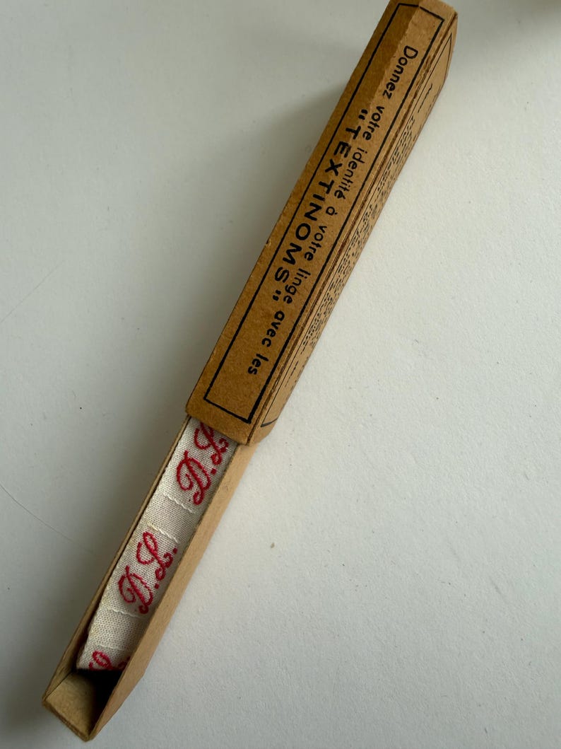 May include: A long, narrow, light brown cardboard box containing a strip of off-white fabric tape with the red embroidered initials D.L. The box lid features black text that reads: Donnez votre identit&eacute; &agrave; votre linge avec les TEXTINOMS.