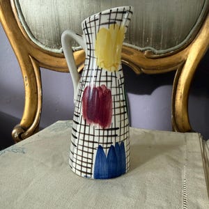 Puede incluir: Una jarra de cerámica con base color crema y un patrón de cuadrícula negra. El exterior está decorado con pinceladas pintadas a mano en amarillo, rojo oscuro y azul. Tiene un asa curva y un diseño de pico cónico y ensanchado.