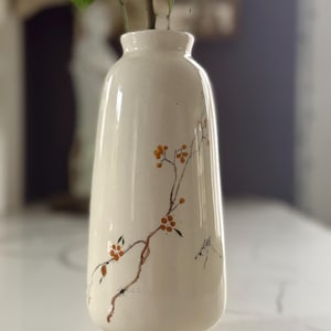 Puede incluir: Jarrón de cerámica color crema con un diseño de rama floral en marrón y naranja. El jarrón tiene un cuerpo redondeado y un cuello estrecho, y sostiene unos tallos de vegetación y pequeñas flores blancas. El jarrón está sobre una superficie blanca.