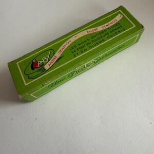 May include: A rectangular green cardboard box with white borders and text. The box features the text ANDRE DEMENOU, LES NOMS ENTIERS TISSES SE FONT EN PLUSIEURS GENRES ET EN DIVERS COLORIS, and Une Grosse Garantie, along with a logo showing a red bird.