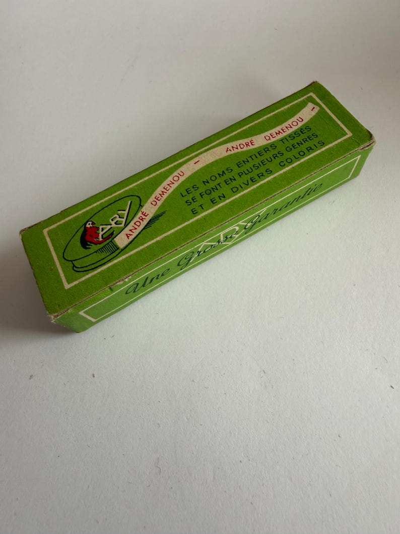 May include: A rectangular green cardboard box with white borders and text. The box features the text ANDRE DEMENOU, LES NOMS ENTIERS TISSES SE FONT EN PLUSIEURS GENRES ET EN DIVERS COLORIS, and Une Grosse Garantie, along with a logo showing a red bird.