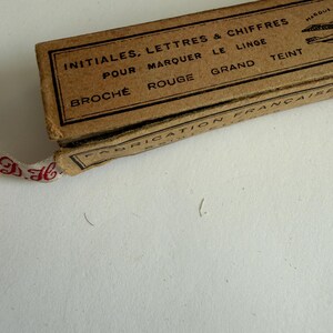 May include: A rectangular brown cardboard box with black printed text reading INITIALES, LETTRES & CHIFFRES POUR MARQUER LE LINGE, BROCH&Eacute; ROUGE GRAND TEINT, FABRICATION FRAN&Ccedil;AISE. A white fabric ribbon with red embroidered initials extends from the box.