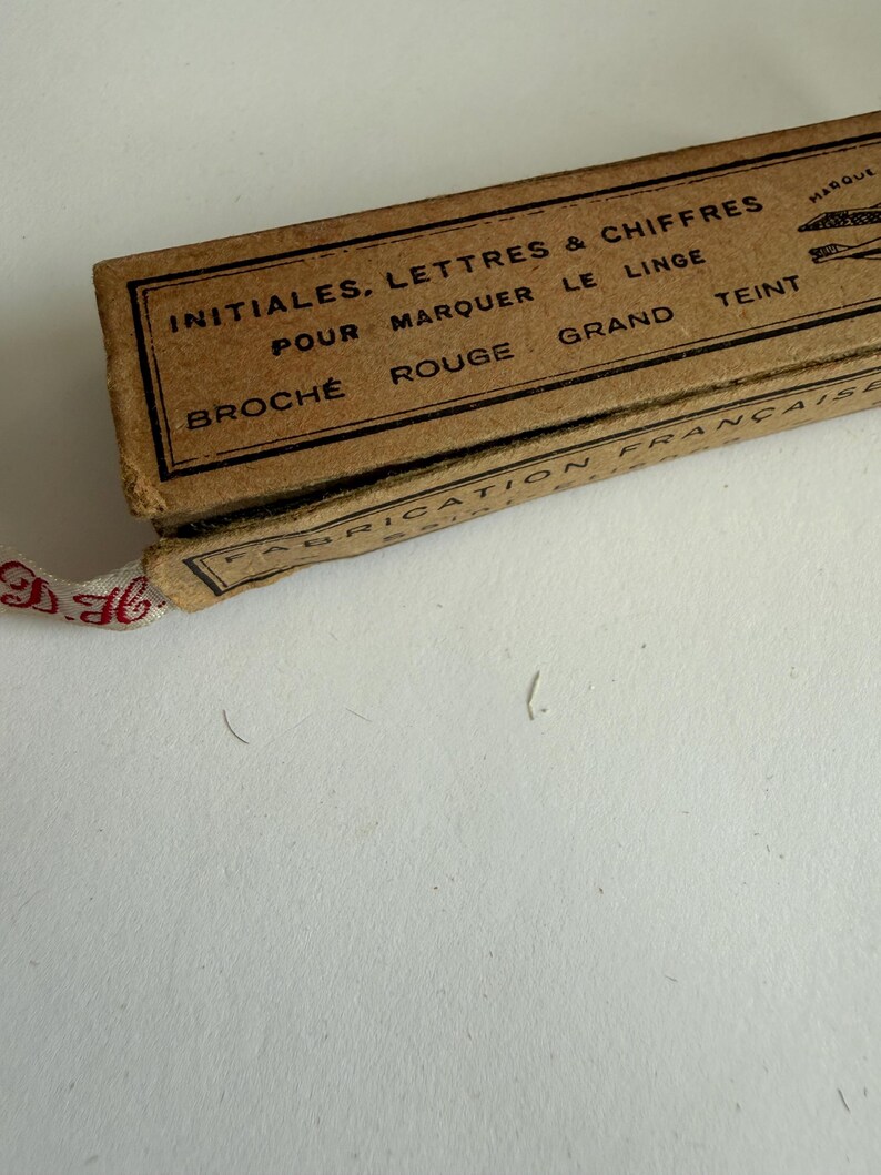 May include: A rectangular brown cardboard box with black printed text reading INITIALES, LETTRES & CHIFFRES POUR MARQUER LE LINGE, BROCH&Eacute; ROUGE GRAND TEINT, FABRICATION FRAN&Ccedil;AISE. A white fabric ribbon with red embroidered initials extends from the box.