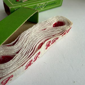 May include: A roll of off-white woven fabric tape featuring repeating red embroidered text that reads R.P. The tape is partially emerging from an open green cardboard box with the text Grosse Garantie printed on the top lid in white.