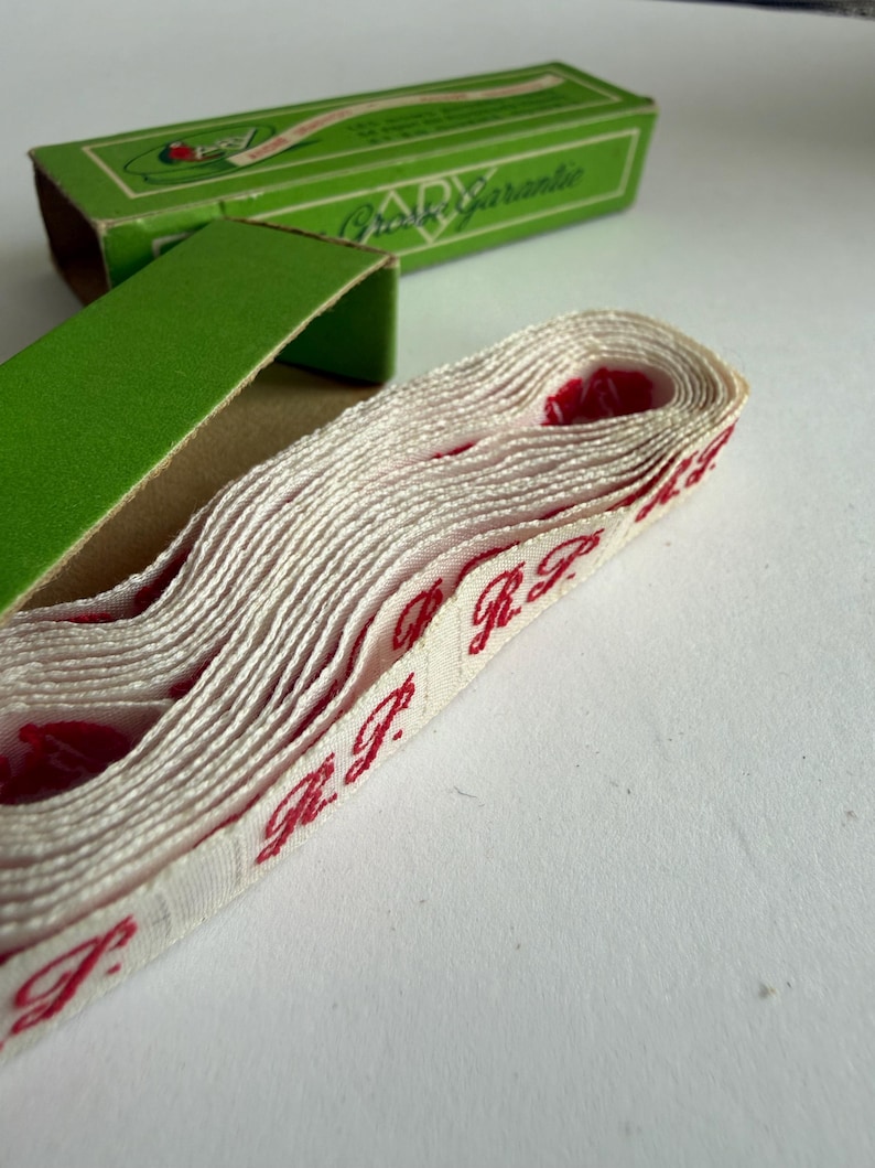 May include: A roll of off-white woven fabric tape featuring repeating red embroidered text that reads R.P. The tape is partially emerging from an open green cardboard box with the text Grosse Garantie printed on the top lid in white.