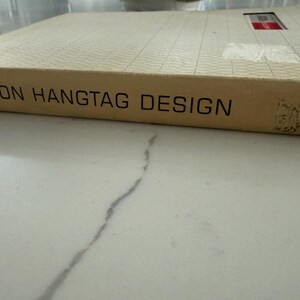 Könnte beinhalten: Ein gebundenes Buch mit dem Titel Fashion Hangtag Design, einem beigen Buchrücken und einem strukturierten, cremefarbenen Einband. Das Buch hat ein kleines rechteckiges Etikett in der oberen rechten Ecke und schwarzen Text.