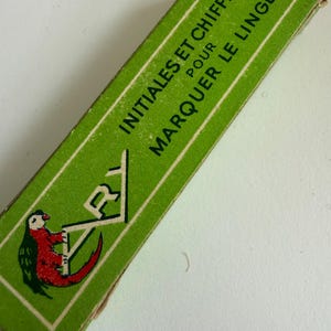 May include: A rectangular green cardboard box featuring the text INITIALES ET CHIFFRES POUR MARQUER LE LINGE in white capital letters. The box displays a graphic illustration of a red and green parrot perched on the letter A.