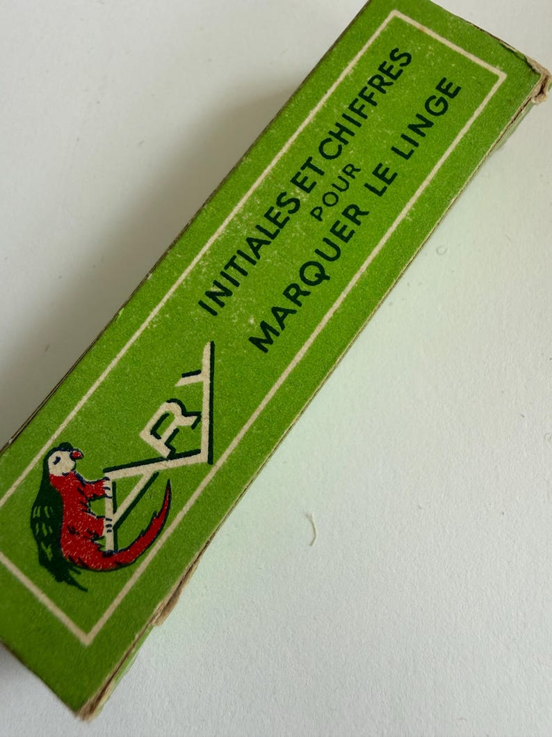 May include: A rectangular green cardboard box featuring the text INITIALES ET CHIFFRES POUR MARQUER LE LINGE in white capital letters. The box displays a graphic illustration of a red and green parrot perched on the letter A.