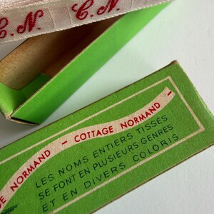 May include: A roll of white woven ribbon with repeating red embroidered initials C.N. sits inside a bright green cardboard box. The box lid features the text COTTAGE NORMAND and LES NOMS ENTIERS TISS&Eacute;S SE FONT EN PLUSIEURS GENRES ET EN DIVERS COLORIS.
