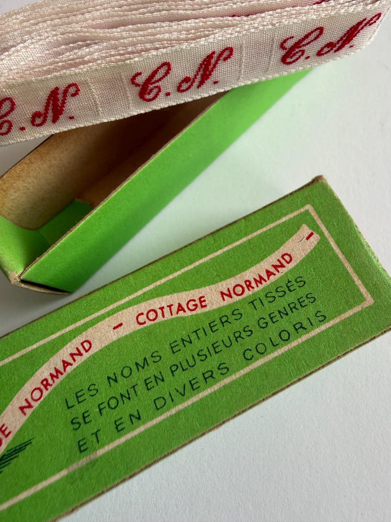 May include: A roll of white woven ribbon with repeating red embroidered initials C.N. sits inside a bright green cardboard box. The box lid features the text COTTAGE NORMAND and LES NOMS ENTIERS TISS&Eacute;S SE FONT EN PLUSIEURS GENRES ET EN DIVERS COLORIS.