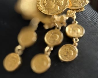 May include: A gold-toned brooch featuring a central circular coin motif with a profile portrait and the text NIKON, surrounded by a decorative wreath. Three dangling chains are attached, each holding smaller circular coin charms with embossed details.