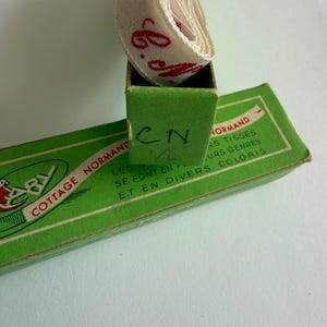May include: A roll of off-white ribbon with red cursive text resting inside a green cardboard box. The box features the text COTTAGE NORMAND, LES RUBANS TISS&Eacute;S SE FONT EN PLUSIEURS GENRES ET EN DIVERS COLORIS, and the handwritten letters CN 1/2.