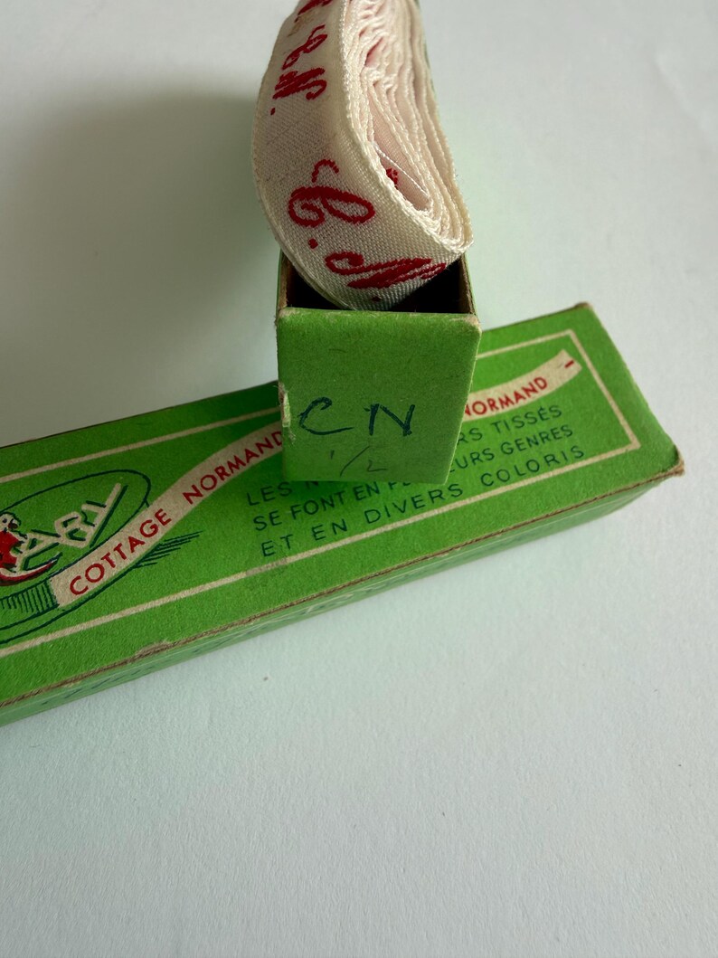 May include: A roll of off-white ribbon with red cursive text resting inside a green cardboard box. The box features the text COTTAGE NORMAND, LES RUBANS TISS&Eacute;S SE FONT EN PLUSIEURS GENRES ET EN DIVERS COLORIS, and the handwritten letters CN 1/2.