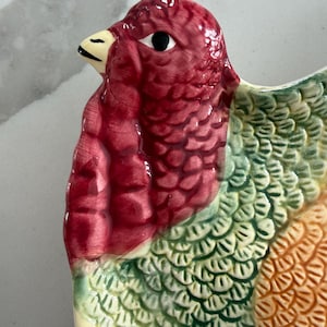 Puede incluir: Un plato de servicio de cerámica con forma de pavo, con un acabado brillante, cabeza y cuello de color rojo intenso, plumas verdes texturizadas y una sección del cuerpo de color naranja claro. El pavo tiene un pequeño pico color crema.