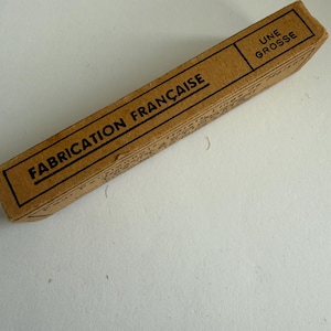 May include: A rectangular brown cardboard box with black text printed on the surface. The text reads "FABRICATION FRAN&Ccedil;AISE" and "UNE GROSSE" in a bold, sans-serif font. The box has a simple, utilitarian design with thin black border lines.