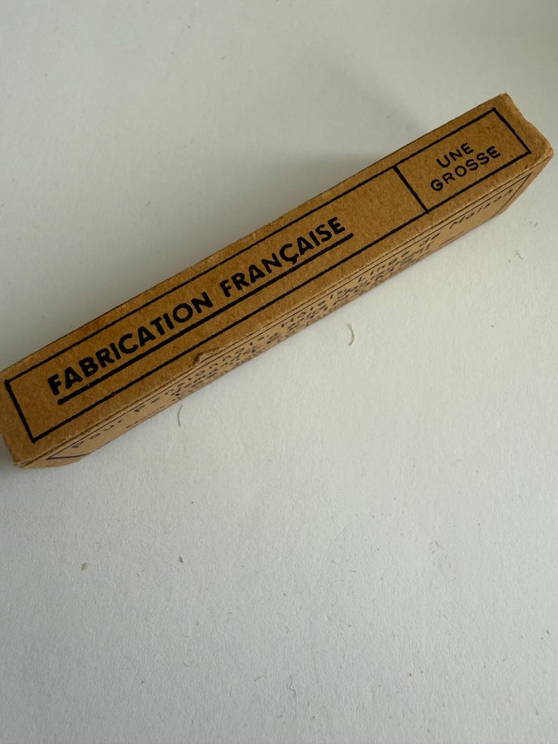 May include: A rectangular brown cardboard box with black text printed on the surface. The text reads "FABRICATION FRAN&Ccedil;AISE" and "UNE GROSSE" in a bold, sans-serif font. The box has a simple, utilitarian design with thin black border lines.