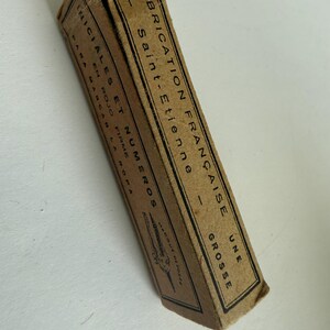 May include: A rectangular tan cardboard box with black text and a small illustration of a tool. The text reads FABRICATION FRAN&Ccedil;AISE, SAINT-ETIENNE, UNE GROSSE, INICIALES ET NUMEROS, EN ROJO FIRME, PARA MARCAR LA ROPA, and MARQUE DEPOSEE.