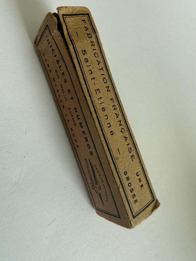 May include: A rectangular tan cardboard box with black text and a small illustration of a tool. The text reads FABRICATION FRAN&Ccedil;AISE, SAINT-ETIENNE, UNE GROSSE, INICIALES ET NUMEROS, EN ROJO FIRME, PARA MARCAR LA ROPA, and MARQUE DEPOSEE.