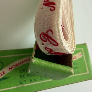 May include: A coiled off-white fabric ribbon with red cursive text reading N.G. emerging from a green cardboard box. The box features printed text including the words NORMAND, LES, SE FU, and ET EN DI in a dark green font on a light green background.