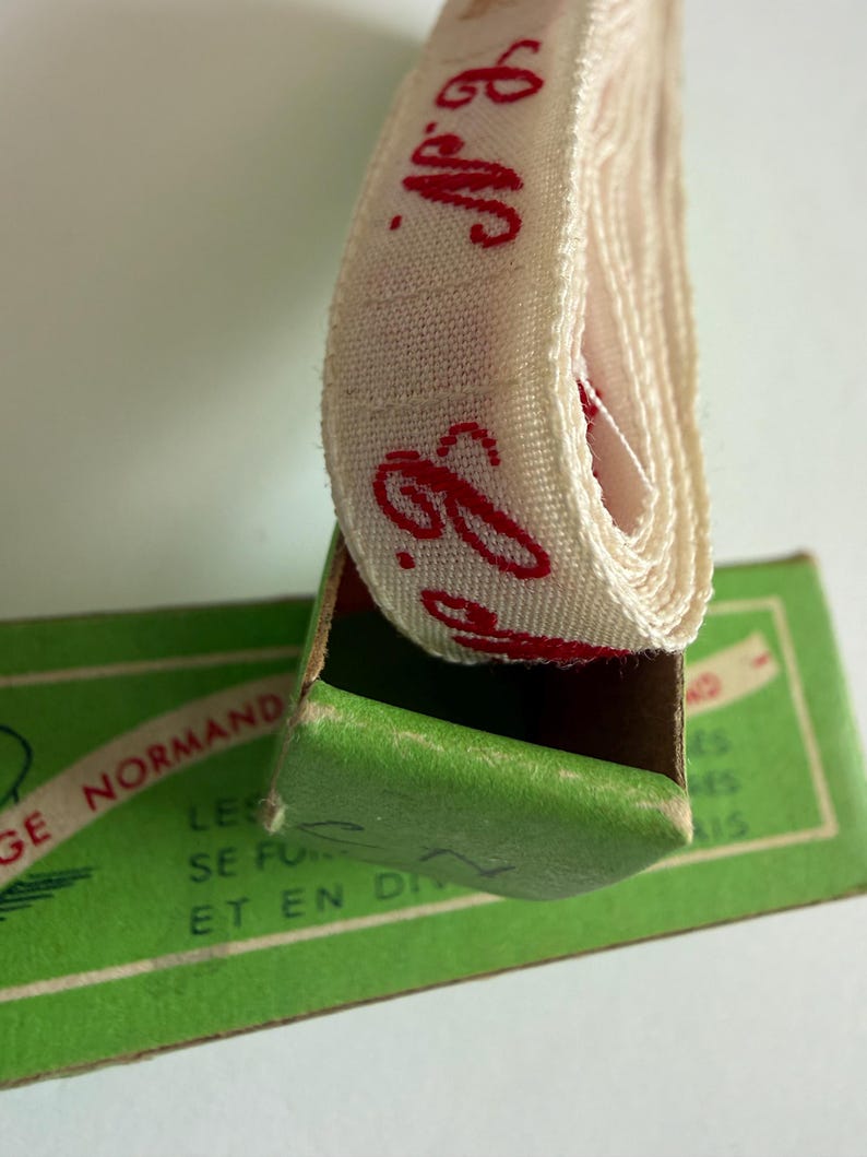 May include: A coiled off-white fabric ribbon with red cursive text reading N.G. emerging from a green cardboard box. The box features printed text including the words NORMAND, LES, SE FU, and ET EN DI in a dark green font on a light green background.