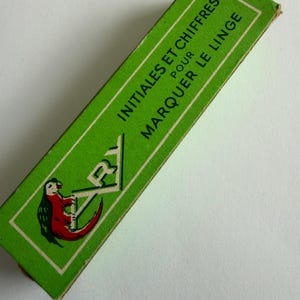 May include: A rectangular green cardboard box featuring the text INITIALES ET CHIFFRES POUR MARQUER LE LINGE in black lettering. The box displays a graphic illustration of a red and green parrot perched on the letters ARY in a white frame.