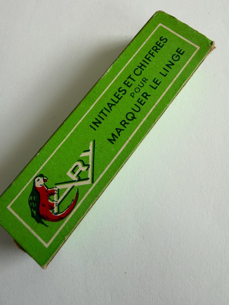May include: A rectangular green cardboard box featuring the text INITIALES ET CHIFFRES POUR MARQUER LE LINGE in black lettering. The box displays a graphic illustration of a red and green parrot perched on the letters ARY in a white frame.
